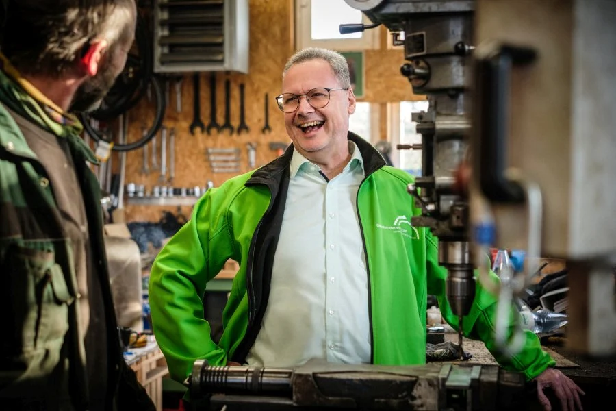 Arne Bär lacht in einer Werkstatt im Gespräch mit einem Handwerker